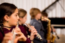 Zwei Kinder spielen nebeneinander Querflöte, dahinter weitere Kinder, eines mit Saxofon